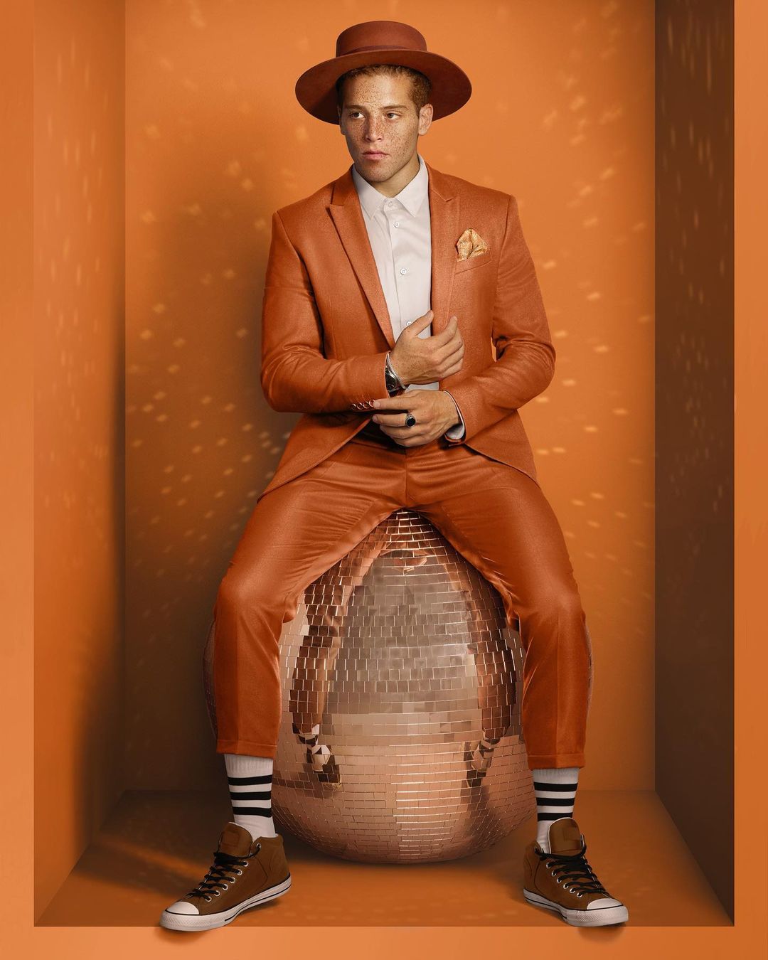 Man in orange suit sitting on disco ball in orange photo studio. He wears a matching hat and brown sneakers with striped socks.